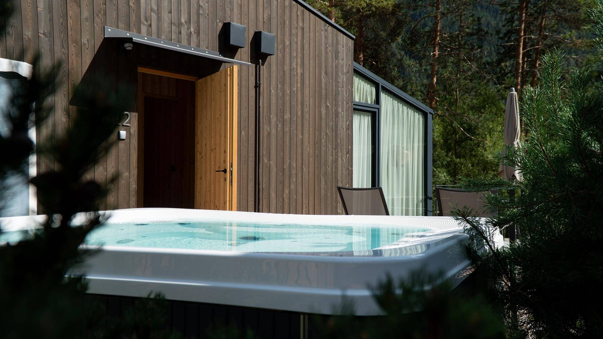 Una vasca idromassaggio all'esterno di una moderna cabina in legno, circondata dal verde e dagli alberi. - Skyview Chalets
