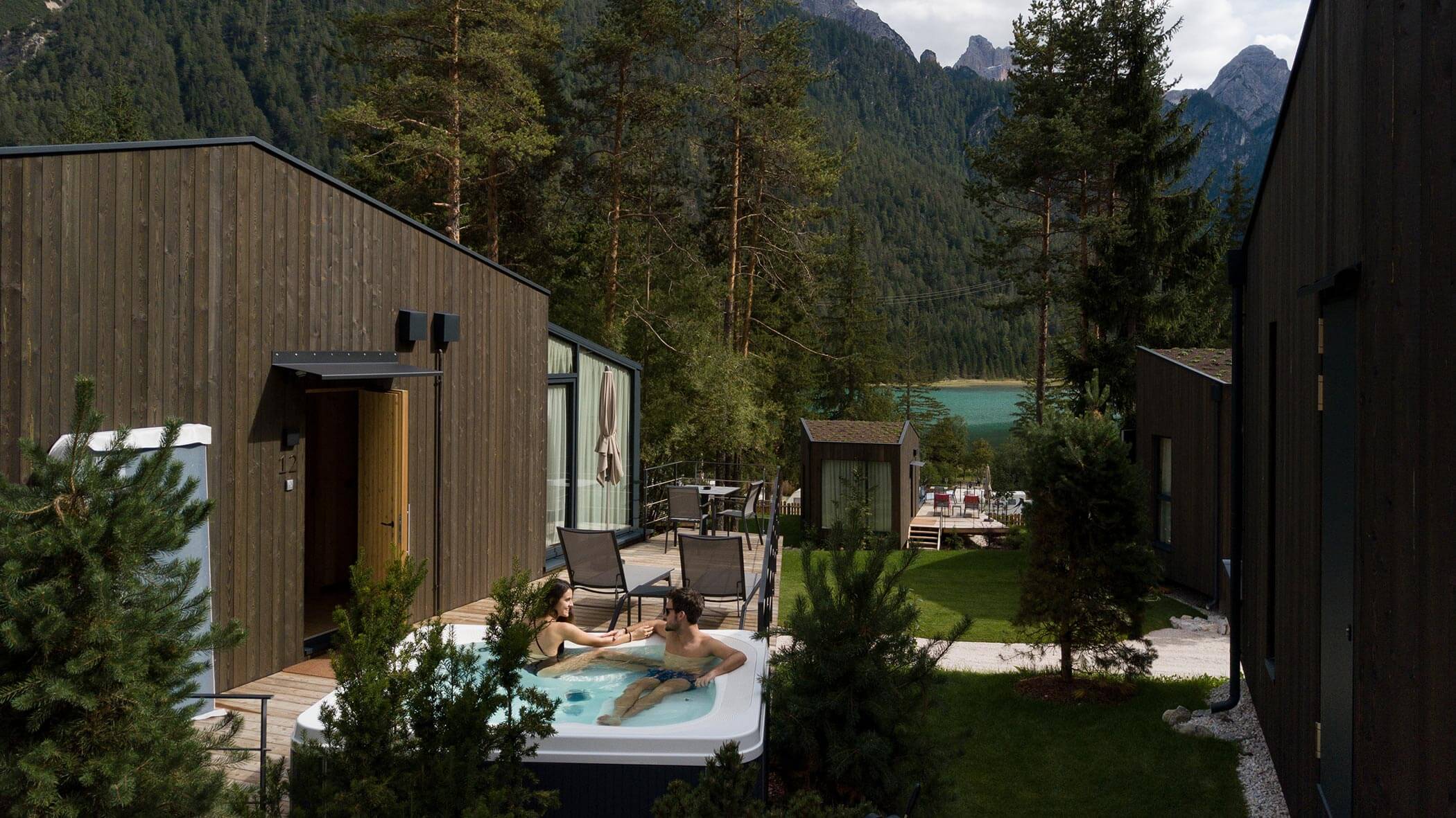 Due persone si rilassano in una vasca idromassaggio all'esterno di moderne cabine, circondate da alberi e montagne vicino a un lago. - Skyview Chalets