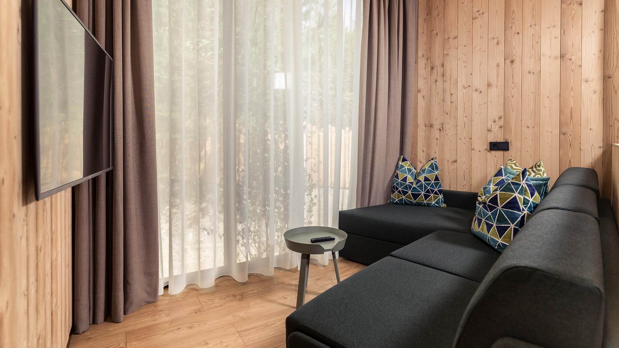 Soggiorno moderno con pareti rivestite in legno, divano grigio, cuscini a fantasia, TV e tende trasparenti. - Skyview Chalets