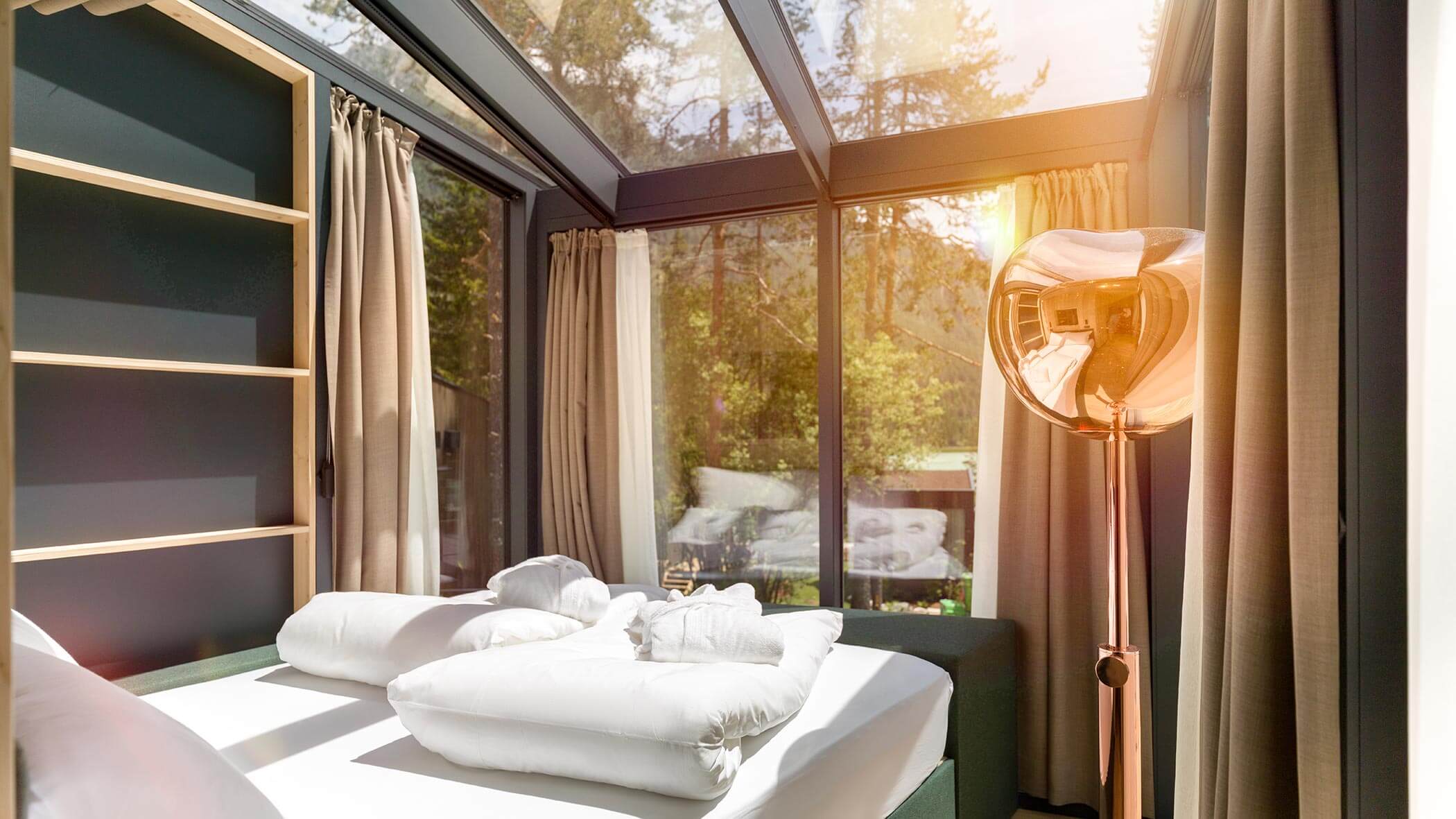 Camera da letto moderna illuminata dal sole con pareti e soffitto in vetro, tende beige e una scultorea lampada da terra metallica. - Skyview Chalets