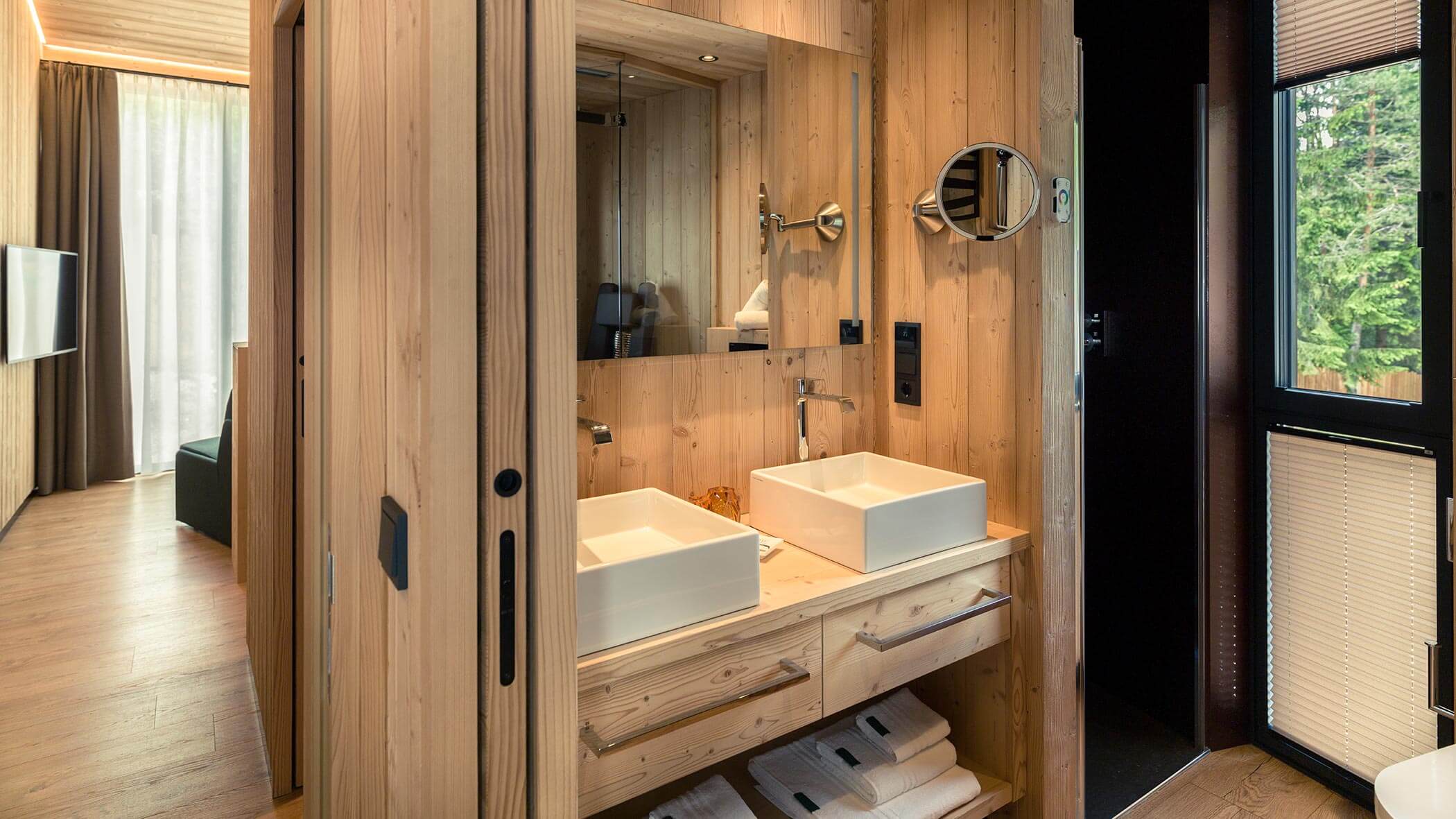 Bagno moderno in legno con doppio lavabo, specchi e asciugamani, accanto a una finestra con vista sulla natura. - Skyview Chalets