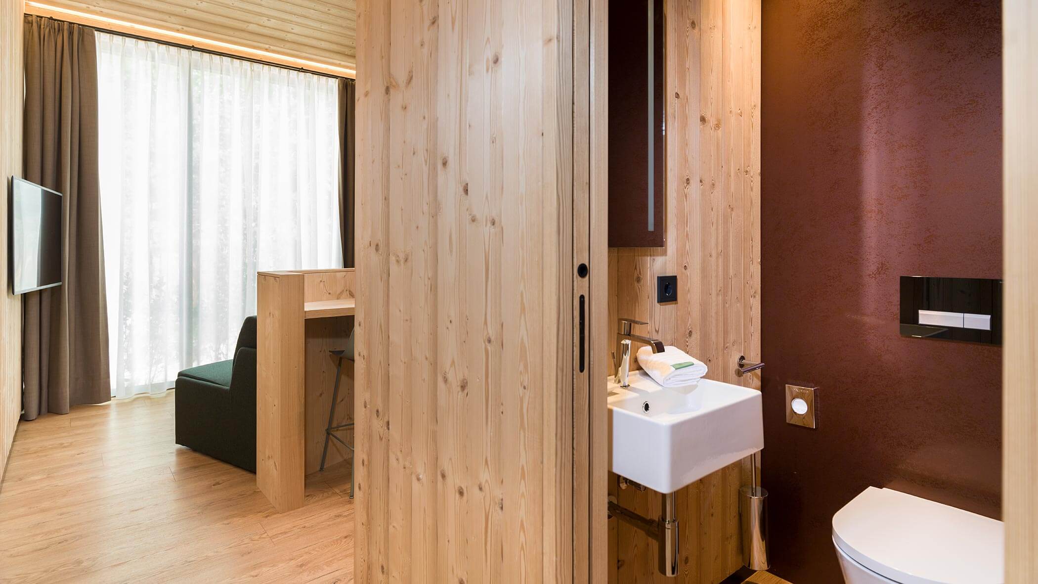Camera d'albergo moderna con pannelli in legno, un piccolo bagno, una scrivania, un divano e un'ampia finestra con tende trasparenti. - Skyview Chalets